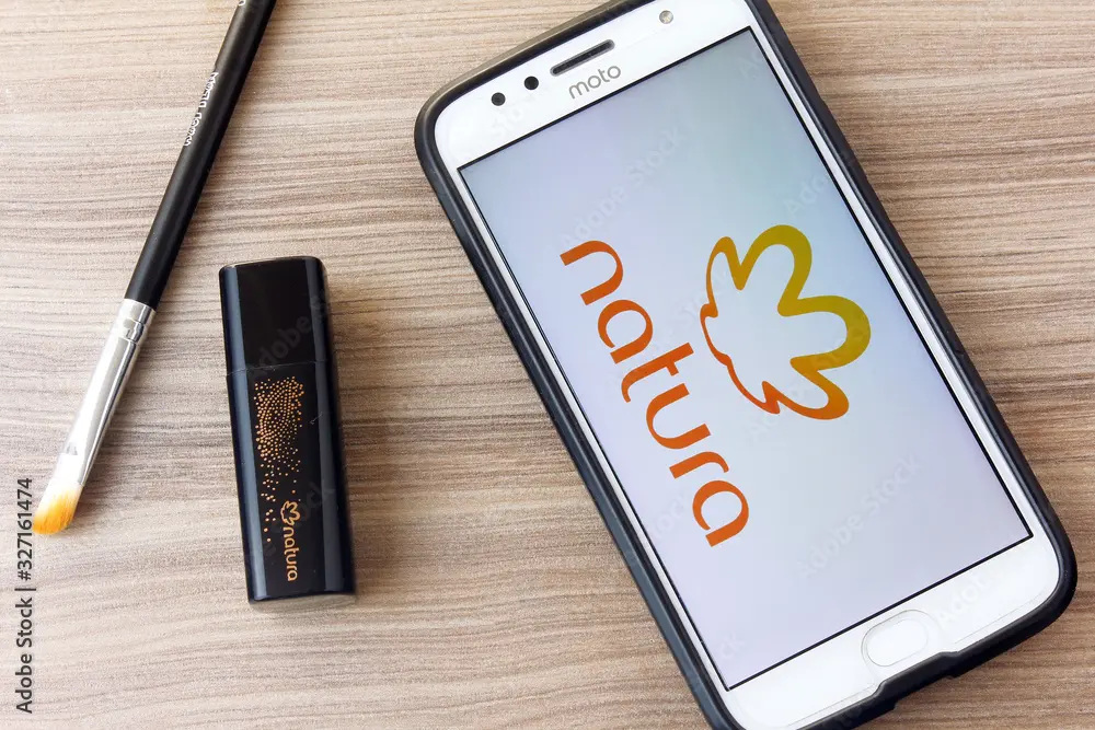 Logo da Natura exibido na tela de um celular, junto com produtos de maquiagem e um pincel.