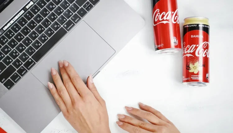 Coca-Cola FEMSA Brasil tem vaga aberta! Veja como garantir um emprego com ótimos benefícios. Pessoa utilizando um laptop com duas latas de Coca-Cola FEMSA sobre a mesa, mostrando o ambiente de trabalho moderno.
