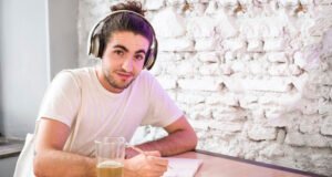 Jovem com coque estudando com fones de ouvido enquanto escuta podcasts para aprender inglês. Copo com líquido amarelado ao lado, fundo da parede branca texturizada.
